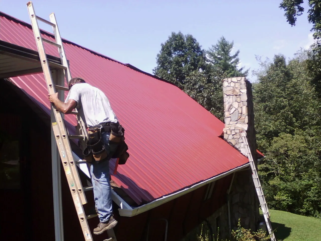 Roofer on a ladder beside a red metal roof and stone chimney — Soffit & Fascia Repair by Roofing Brothers in Tensed